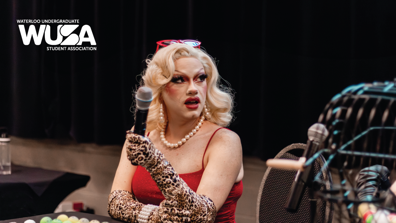 A person in a glamorous outfit holds a microphone at an all-ages event, adorned with the "Waterloo Undergraduate Student Association" logo.