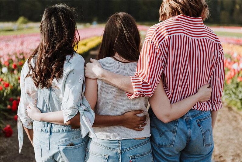 Three people with their arms around each other stand in a field of flowers, viewed from behind, illustrating the comforting warmth and support.