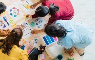 A Marketing Coordinator joins a team collaborating on a creative project. Around the table, they dive into colorful materials, papers, and a laptop, ensuring every detail shines in their SEO strategy for Winter 2025.