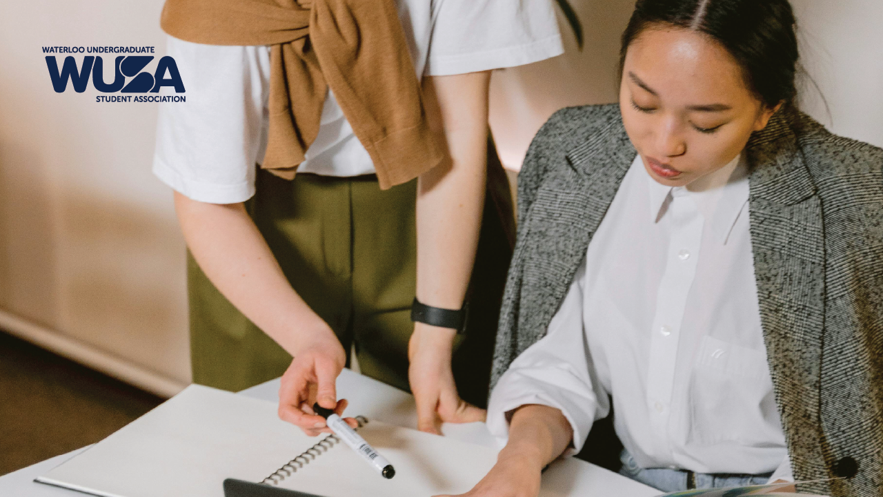 Two people collaborating at a roundtable with a notebook, pen, and a laptop.