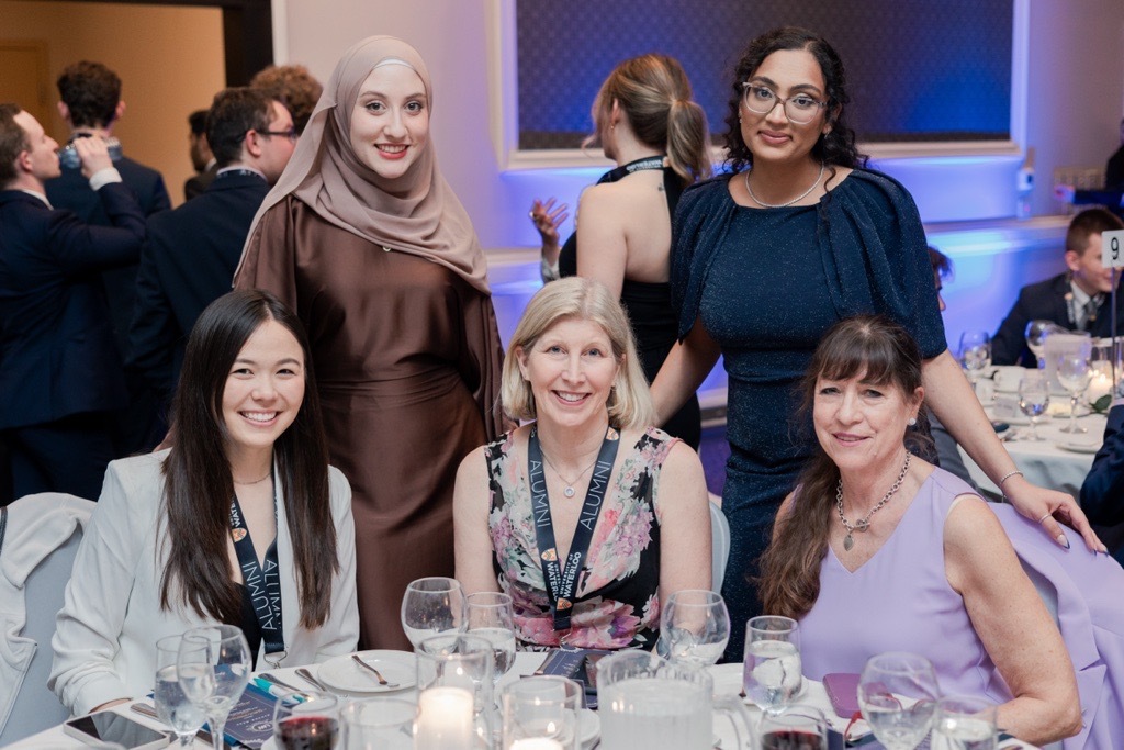 Five women smiling at a gala, seated and standing around a candlelit table with glasses, as they celebrate their achievements in the aviation industry.