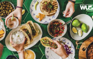 Hands eagerly reaching for Taco Tuesday delights—tacos, salsa, and drinks spread across a vibrant green table. Lime and olives add a fresh touch to the scene. Logo elegantly placed in the top right corner.