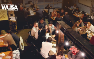 A bustling restaurant with people eating and chatting at tables, highlighted by the Waterloo Undergraduate Student Association logo, hosts a lively trivia night inspired by Abe Erb in Uptown Waterloo.