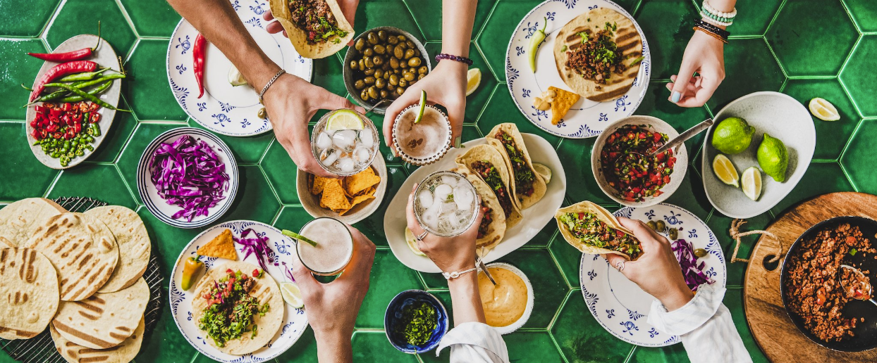 Hands reaching for tacos and drinks on a green tiled table, embodying the spirit of Taco Tuesday, surrounded by colorful ingredients and garnishes.