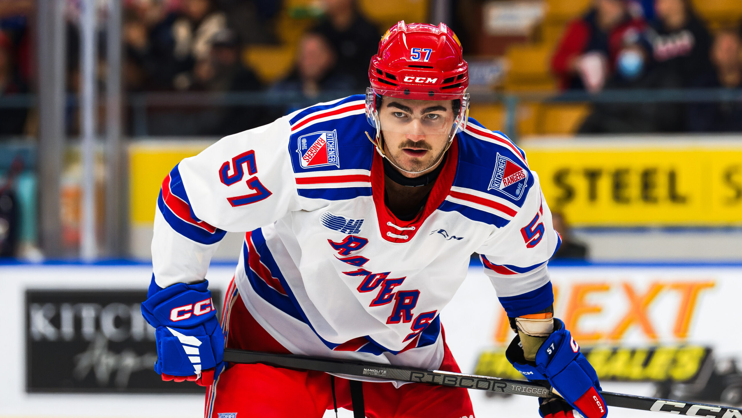 A Ranger in a blue, white, and red uniform stands on the ice with intense focus, ready for Game Night, clutching his stick with determination.