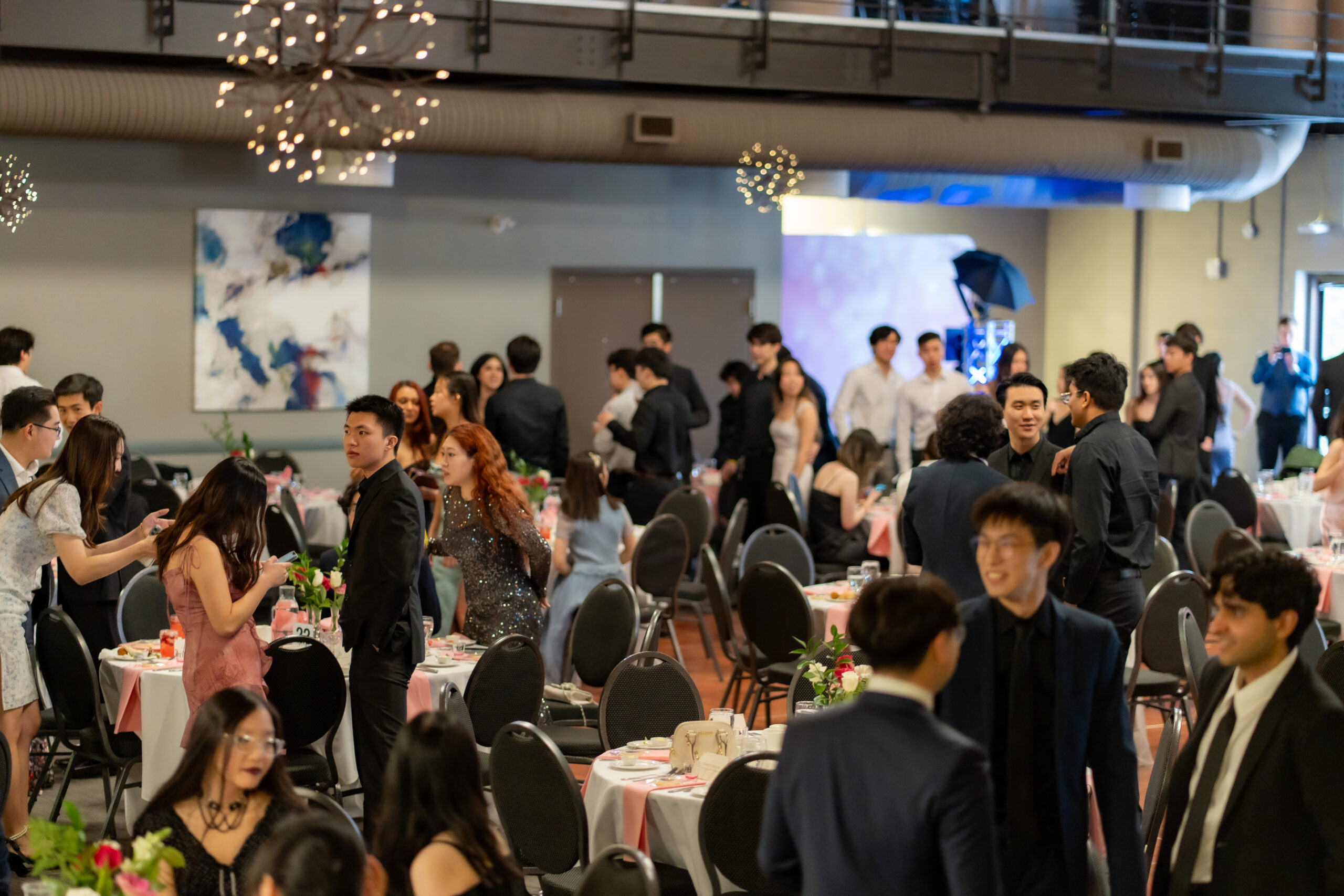 A large group of people socialize in a decorated event space with round tables and chairs.