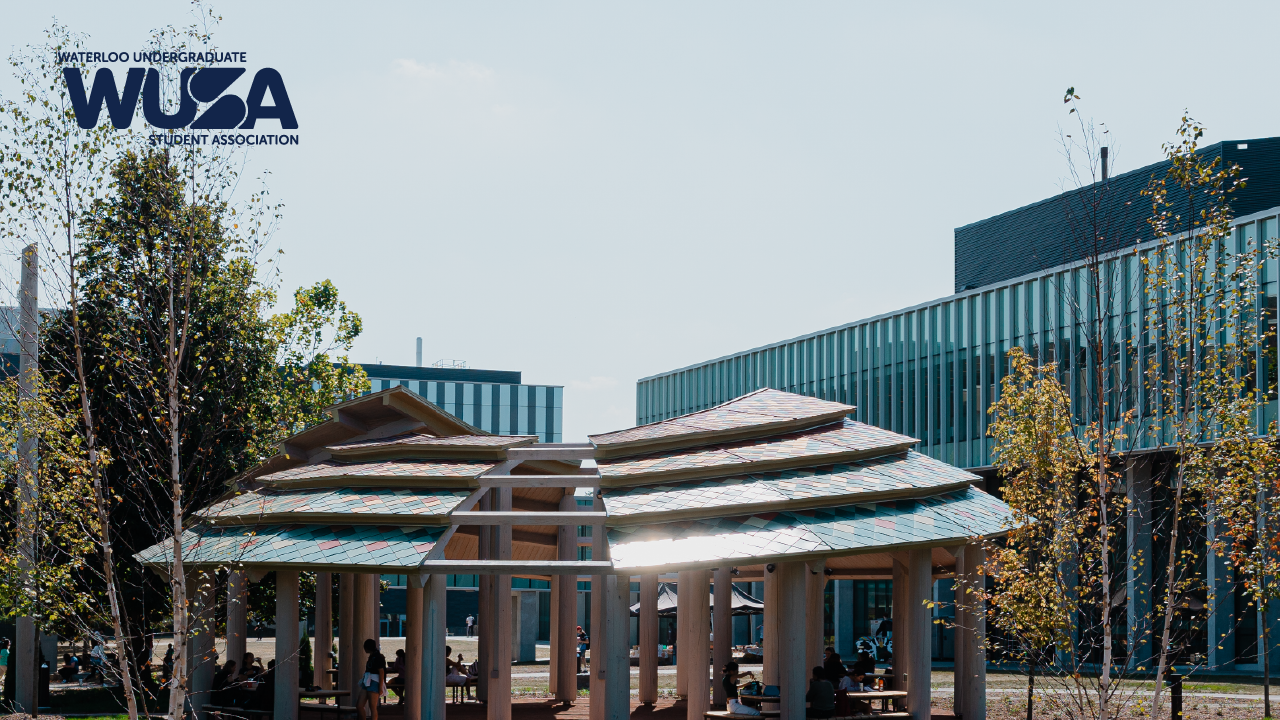 An outdoor wooden pavilion with a curved roof sits gracefully under a clear sky, surrounded by modern buildings and trees. It serves as a vibrant space for conversation circles, inviting participants to engage deeply in sharing ideas and student feedback.