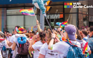 People joyfully celebrate Pride at the Toronto Parade, waving rainbow flags and holding plush toys, as the "WUSA Glow Centre" sign shines in the background.