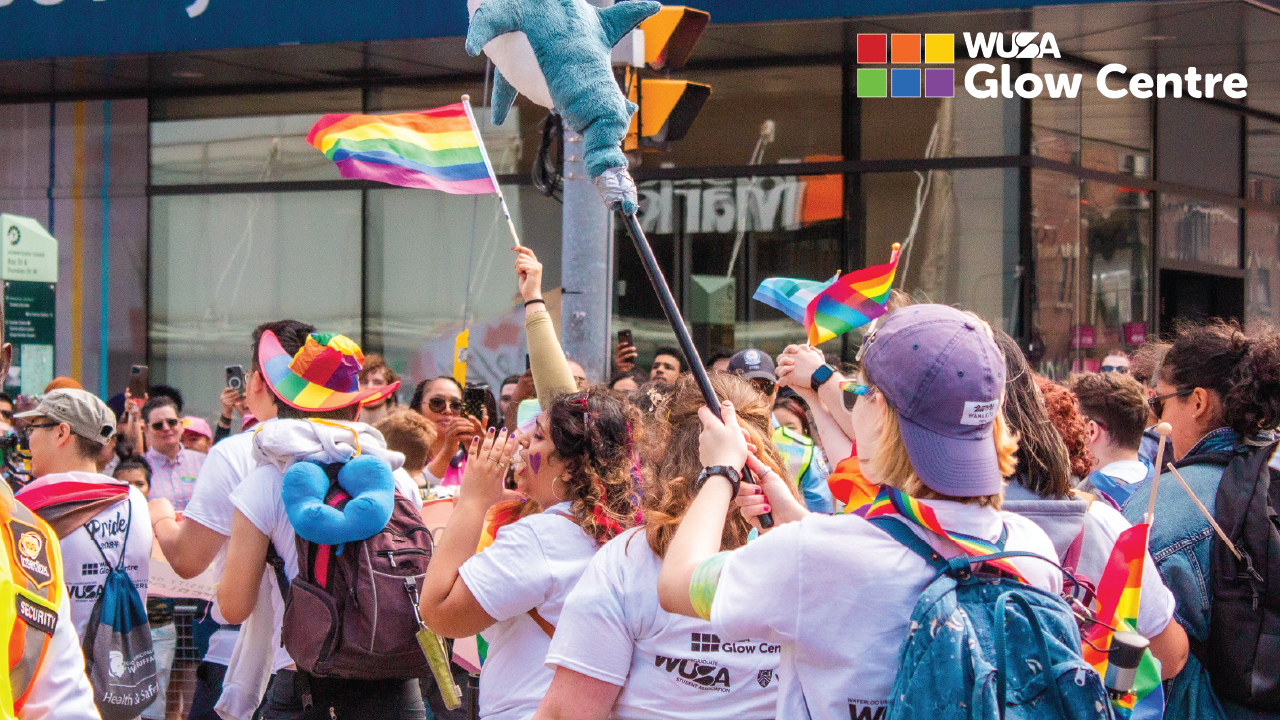 People joyfully celebrate Pride at the Toronto Parade, waving rainbow flags and holding plush toys, as the "WUSA Glow Centre" sign shines in the background.