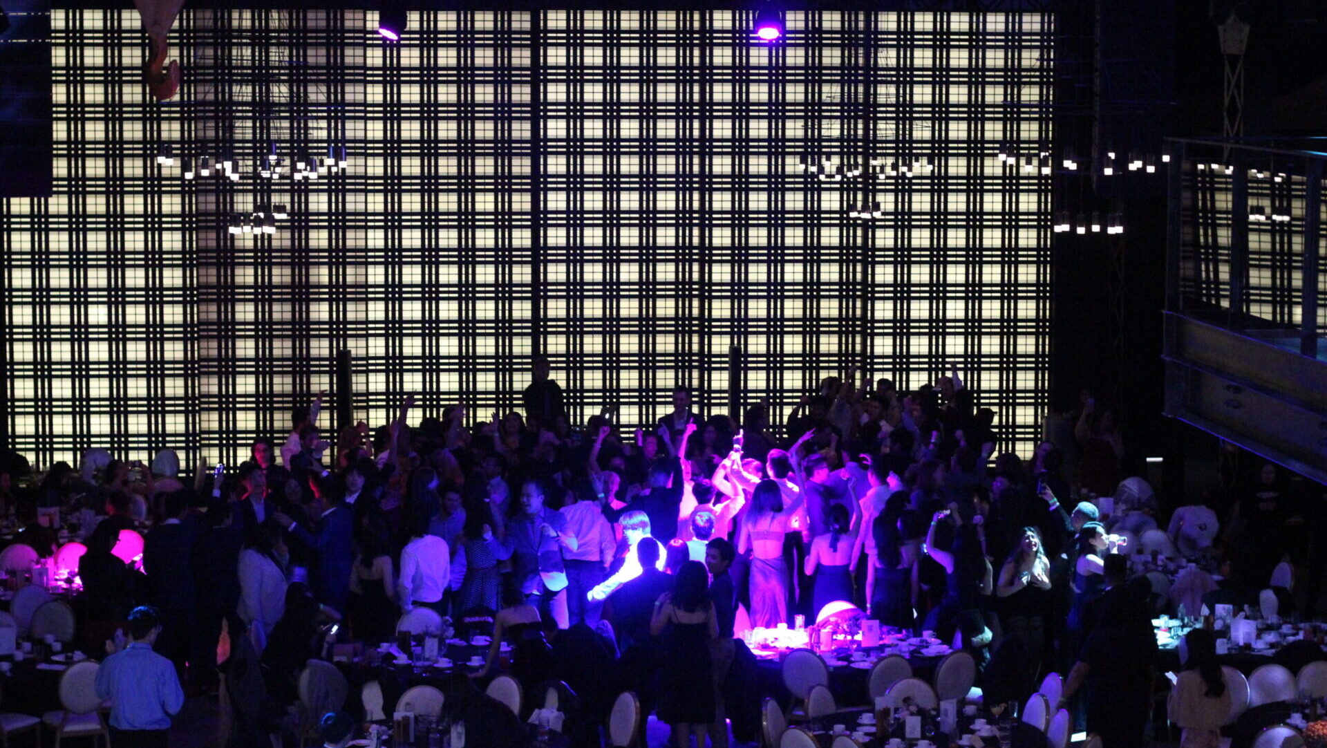 A lively crowd dances in a dimly lit venue, celebrating the Math Grad Ball, as a large checkered light display casts mesmerizing patterns in the background.