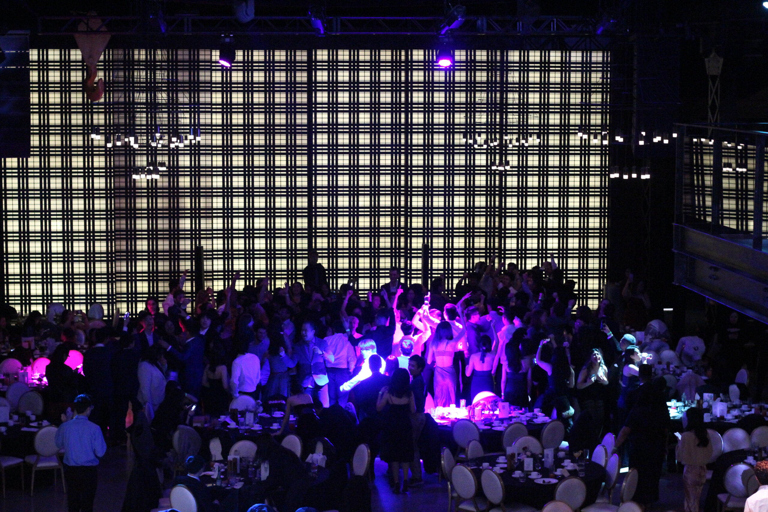 A lively crowd dances in a dimly lit venue, celebrating the Math Grad Ball, as a large checkered light display casts mesmerizing patterns in the background.