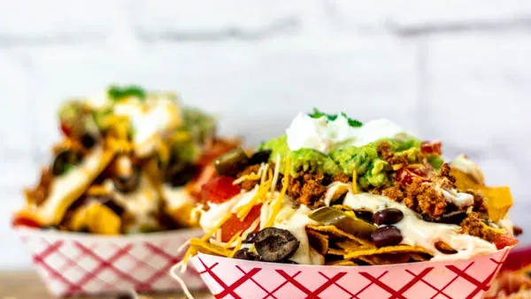 Bowls of loaded nachos with cheese, olives, guacamole, sour cream, and toppings in red checkered containers.