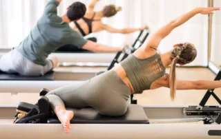 Two people enjoying a Pilates Power Hour on reformer machines, with arms extended overhead.