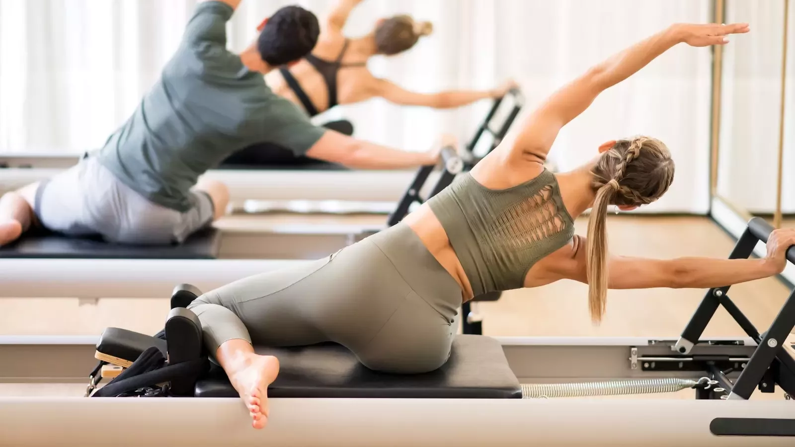 Two people enjoying a Pilates Power Hour on reformer machines, with arms extended overhead.