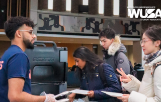 Students mingling at a booth in the Kitchener Campus lobby, with one flashing a peace sign, as they discuss plans for brunch.