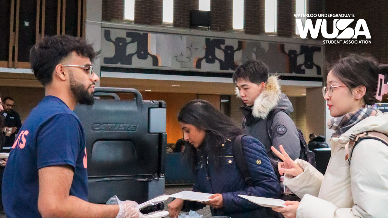 Students mingling at a booth in the Kitchener Campus lobby, with one flashing a peace sign, as they discuss plans for brunch.