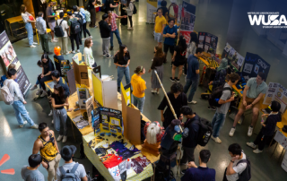 Aerial view of a bustling WUSA club fair with students visiting various booths in a large indoor space, eagerly nominating peers for the upcoming Volunteer Appreciation Awards.