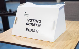 A white voting screen labeled "Elections Ontario" sits on a wooden table, ready for the Ontario Provincial Election.