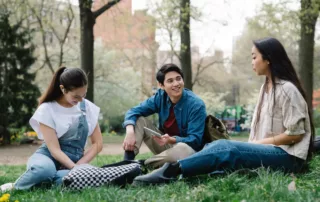 Three people are sitting on the grass in a park, engaged in lively conversation about campus safety, framed by trees and distant buildings.