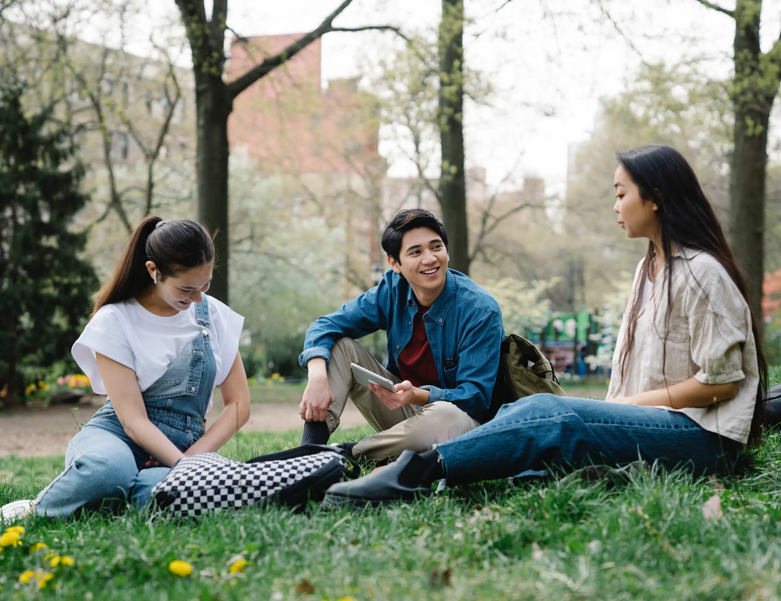 Three people are sitting on the grass in a park, engaged in lively conversation about campus safety, framed by trees and distant buildings.