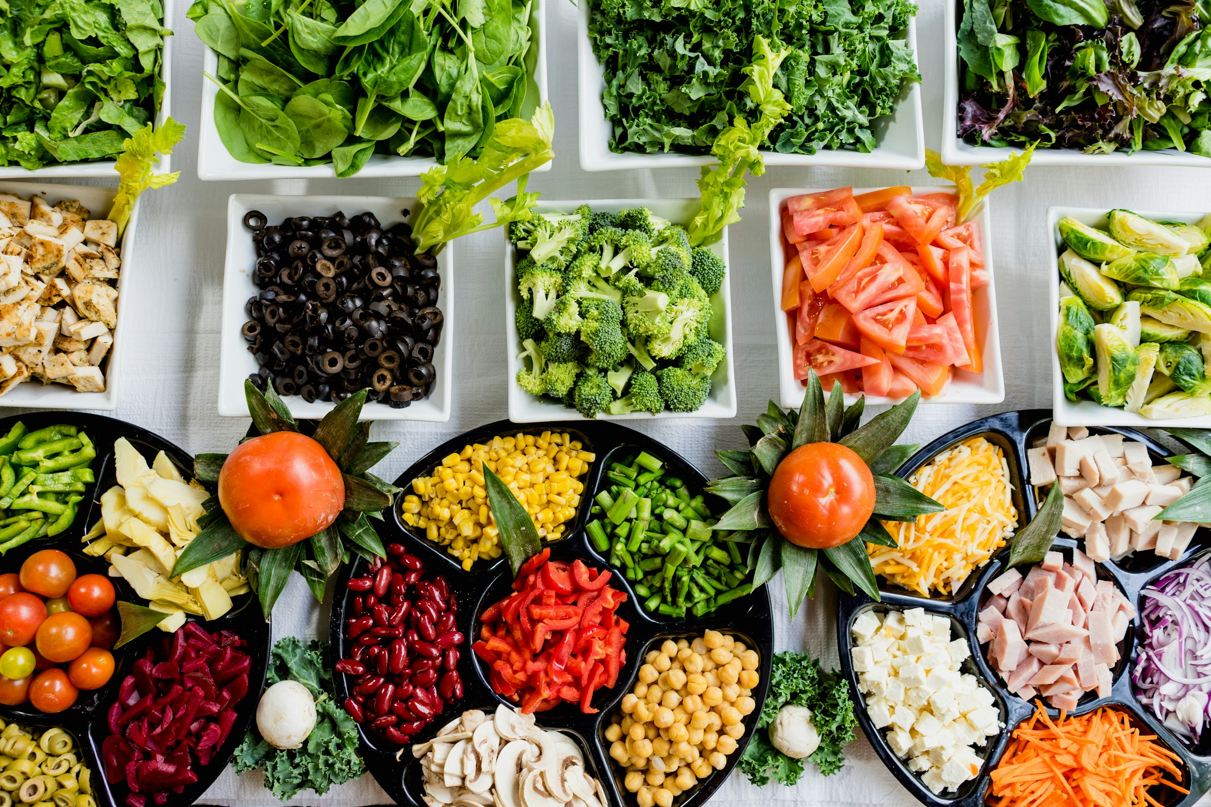 A variety of fresh vegetables and salad ingredients, showcasing a delightful blend of Tastes From Home and Global Kitchen, are meticulously arranged in bowls and trays on a pristine white table.