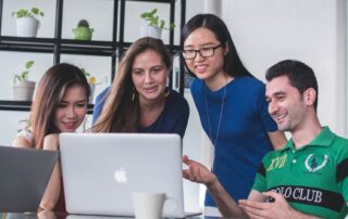 Four international students gathered around a laptop, discussing and smiling in a modern office with plants on shelves.