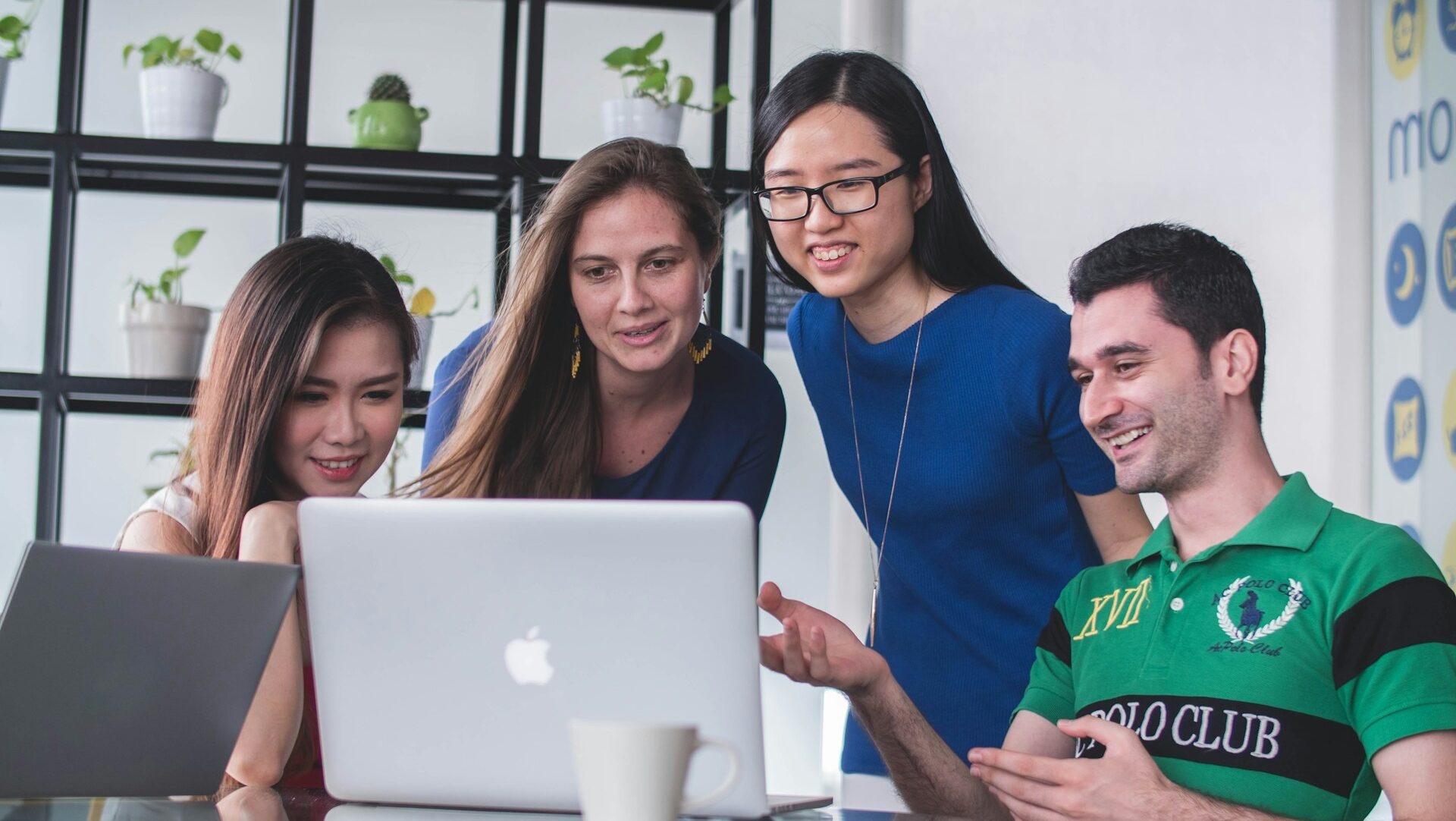 Four international students gathered around a laptop, discussing and smiling in a modern office with plants on shelves.