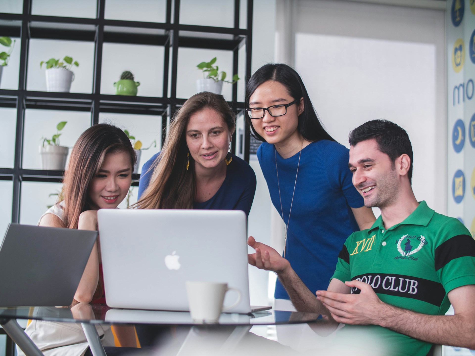 Four international students gathered around a laptop, discussing and smiling in a modern office with plants on shelves.