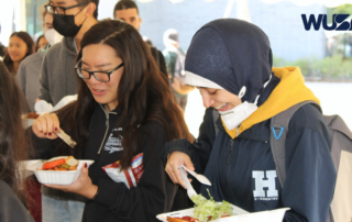 People in line with food trays, smiling and eating, under a canopy with a WUSA logo in the corner.