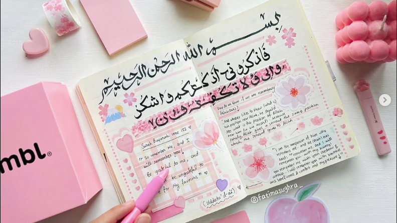 An open journal with a pink aesthetic comes to life under the soft glow of night, featuring elegant handwritten Arabic calligraphy. Cherry blossoms grace the pages as a hand delicately holds a pink pen, capturing an intimate event of artistic expression.