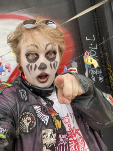 Person with face paint and patches on jacket points at camera.