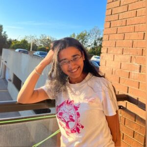 Smiling woman with long hair and glasses leans on a balcony railing in the sunlight, wearing a white t-shirt.