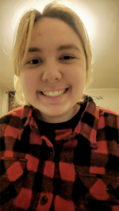 A light-haired person smiling at the camera in a red and black plaid shirt.
