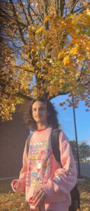 Young person with curly hair wearing a pink graphic sweatshirt stands outside under autumn leaves, holding a drink.