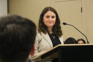 A woman stands at a podium, speaking into a microphone during the Winter 2025 Volunteer Awards, celebrating Student Excellence at a WUSA-hosted indoor event.