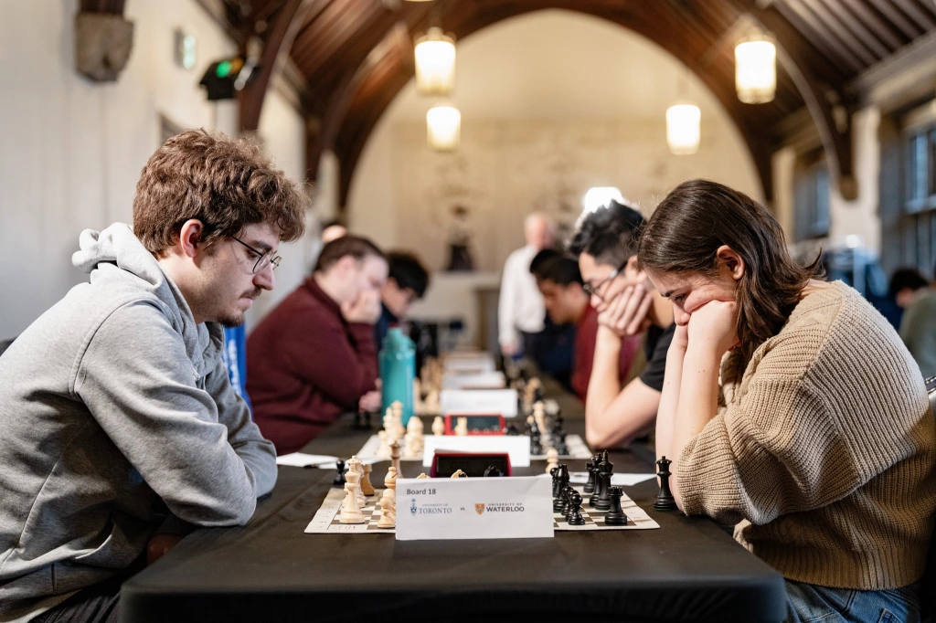 Two people play chess in a focused tournament setting with others competing in the background.