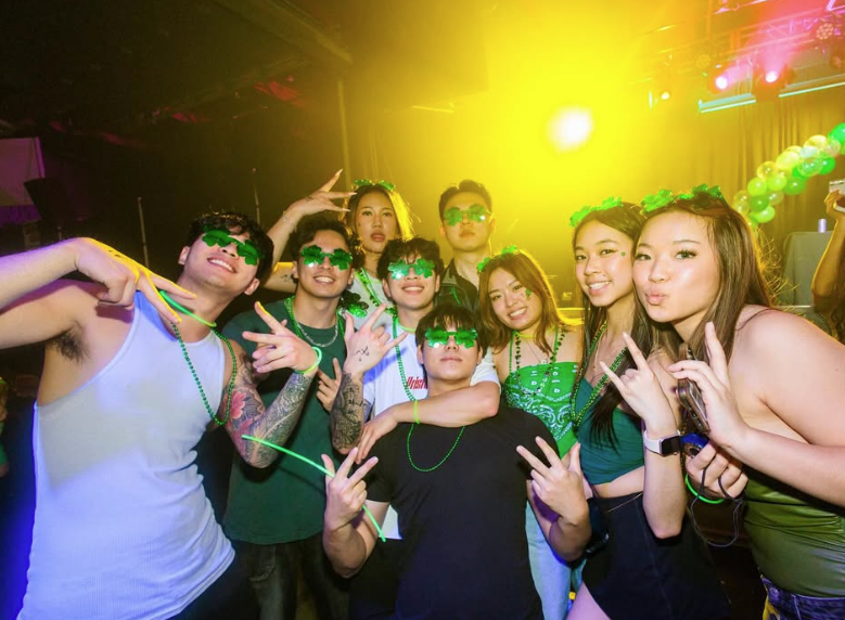 393043da-1a33-45d5-82e7-b99223b0793a A group of young adults in green outfits and accessories pose together at a lively party with bright lighting.