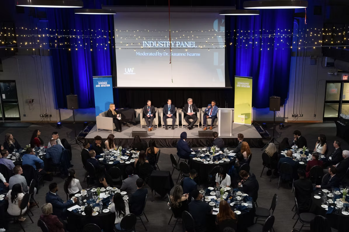 A panel of five speakers sits on stage at an industry event with an audience seated at round tables.
