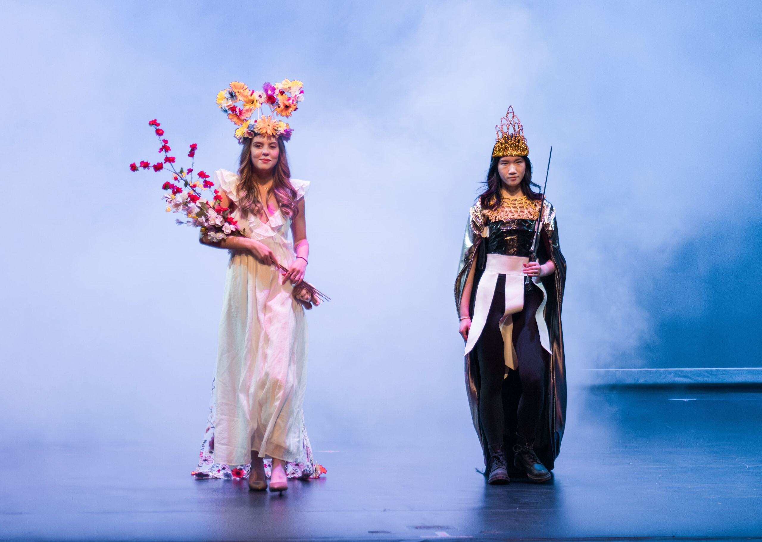 6e495486-e4fe-4f89-9e2b-b852fc729783 Two women in elaborate costumes and crowns stand on stage with a misty, blue-lit background.