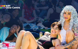Two smiling people sit on grass at a vibrant Tri-Pride outdoor gathering, glowing with joy as more people socialize in the background.