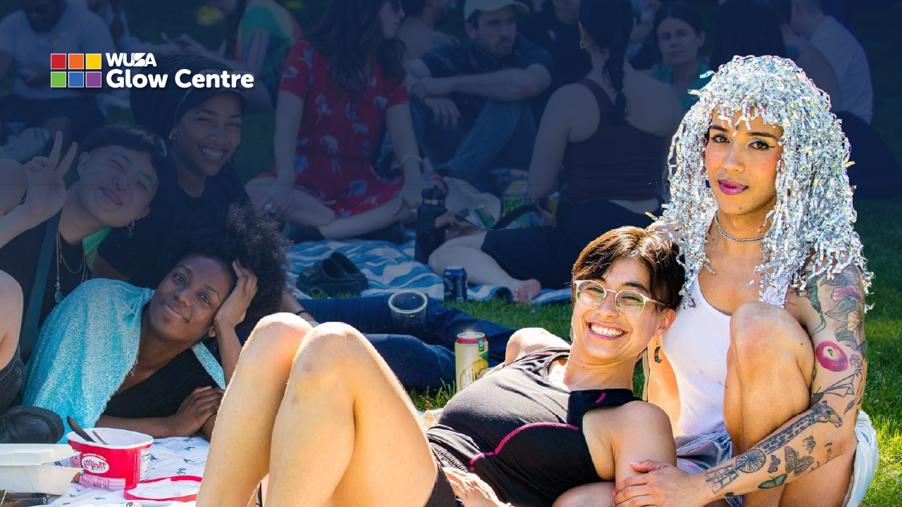 Two smiling people sit on grass at a vibrant Tri-Pride outdoor gathering, glowing with joy as more people socialize in the background.