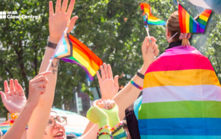 People celebrate pride outdoors at an LGBTQ+ fair, waving rainbow flags and wearing colorful outfits, with trees in the background.