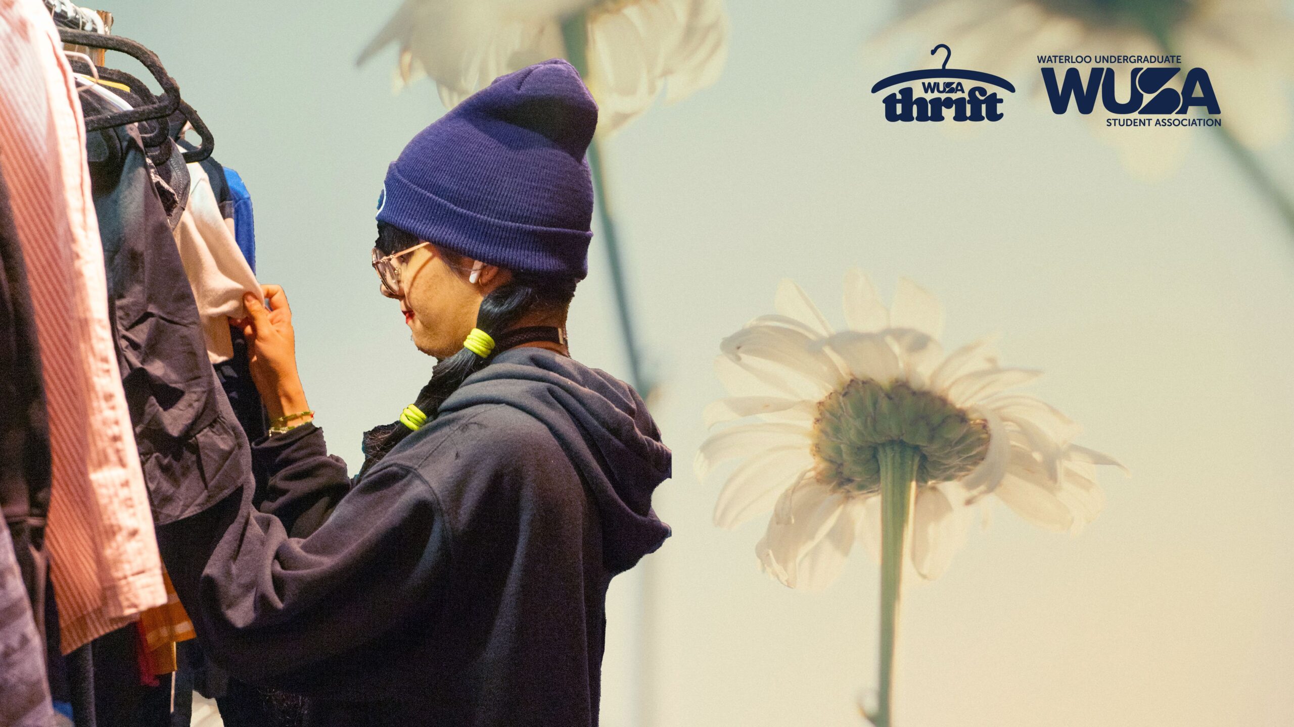 A person in a beanie browses clothes on a rack at the Sip & Thrift Sidewalk Sale, with white flowers and logos in the background.