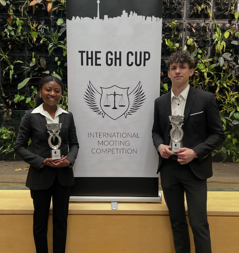 c1c55244-37d7-4f2b-a4a4-fe258ce71934 Two people in suits hold trophies in front of a banner for "The GH Cup International Mooting Competition.