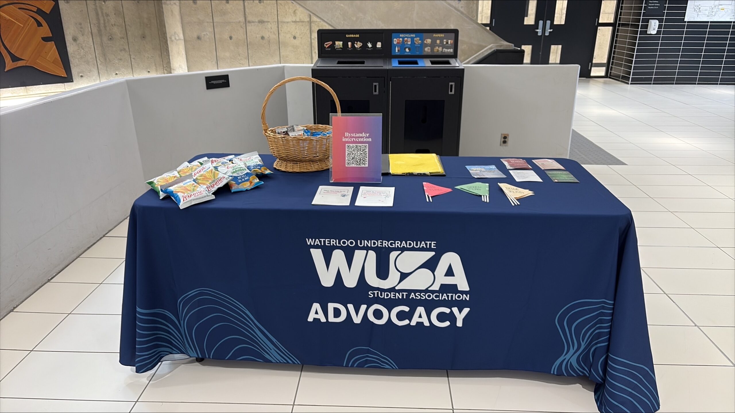 A table with snacks, pamphlets, and signs, covered by a WUSA Advocacy cloth in a building lobby, raises awareness for Sexual Violence Month.