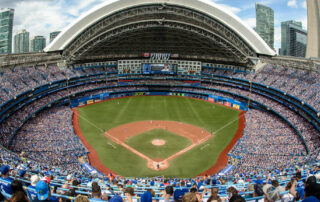 A packed baseball stadium with a retractable roof, fans wearing blue, and a Blue Jays game in progress on the field.