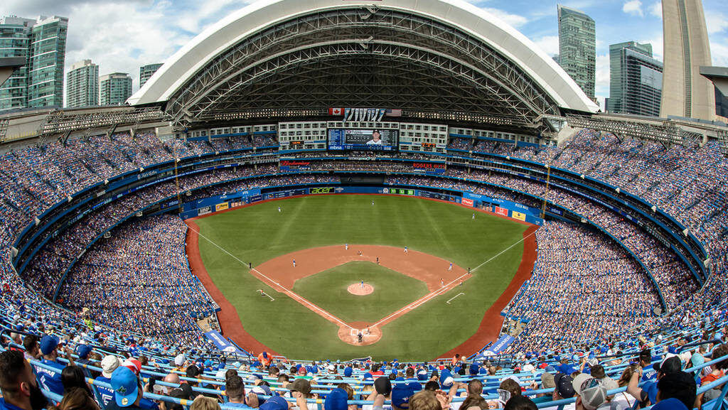 A packed baseball stadium with a retractable roof, fans wearing blue, and a Blue Jays game in progress on the field.