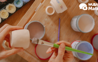 Hands painting a small ceramic pot in a Pot Painting session with brushes and paint jars on a wooden table; WUSA Mates logo in the corner.