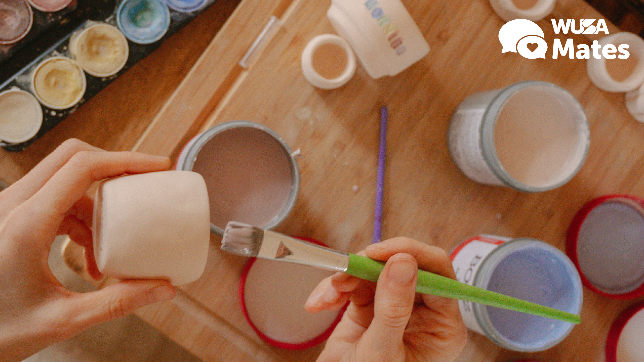 Hands painting a small ceramic pot in a Pot Painting session with brushes and paint jars on a wooden table; WUSA Mates logo in the corner.