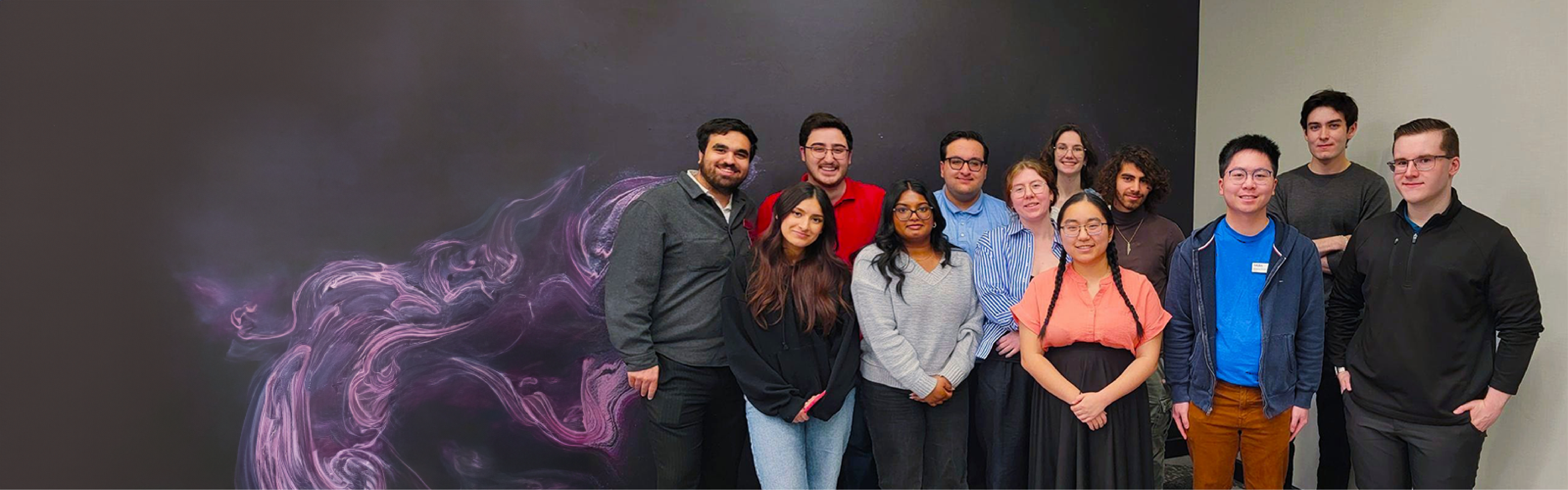 A group of thirteen people, known as Student Bodies, standing and smiling in front of a dark, swirly abstract mural.